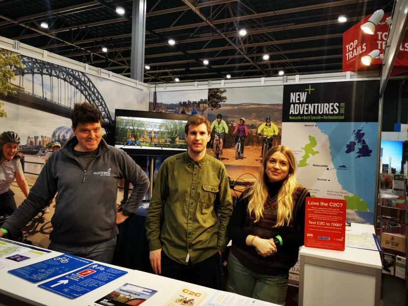 The team of three people on their stand at one of the expos with imagery of the region and cycling in the background.