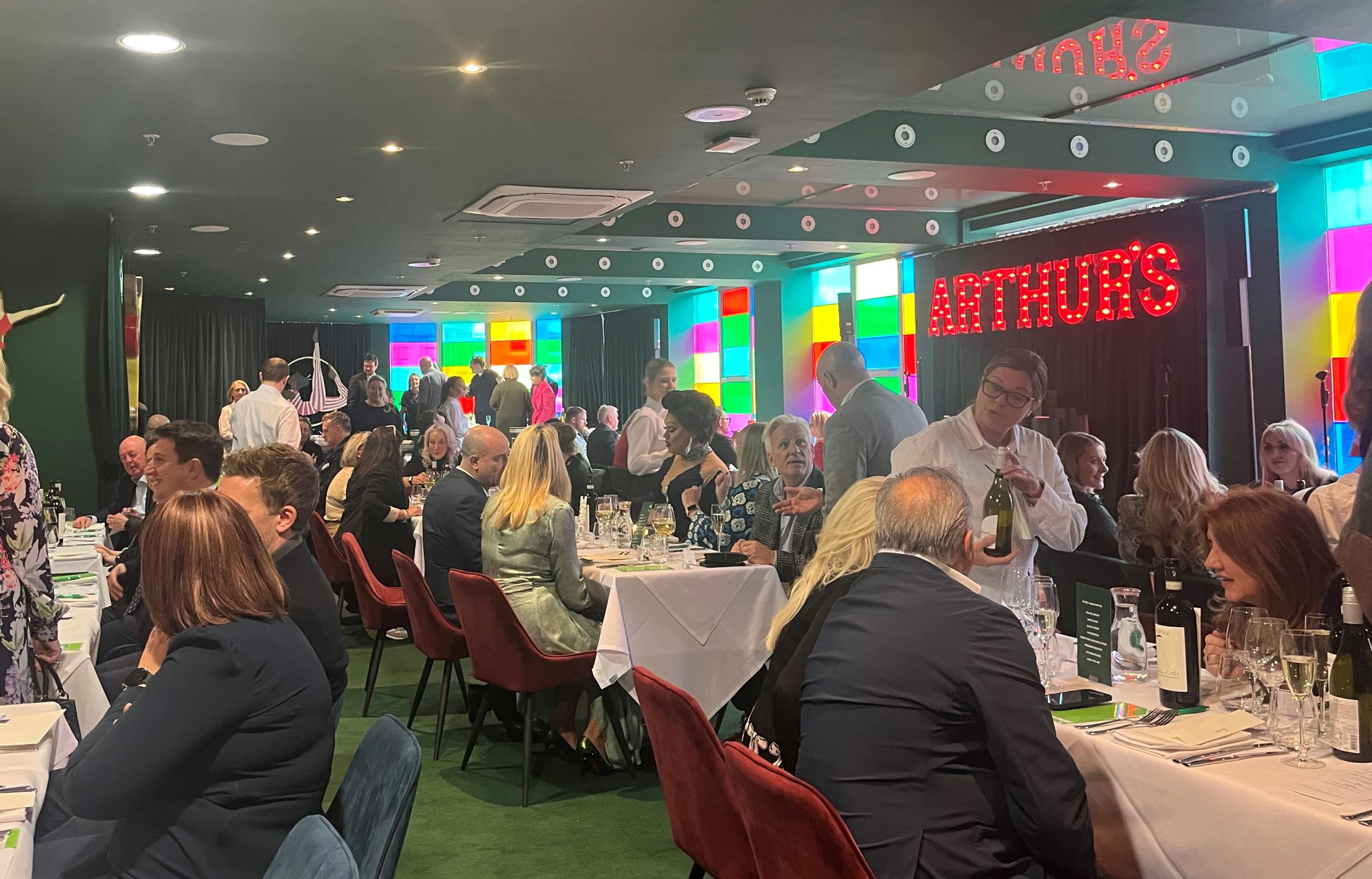 People sat at rows of tables with white tablecloths. The room has green walls and carpets and people are all talking to each other whilst being served by waiters