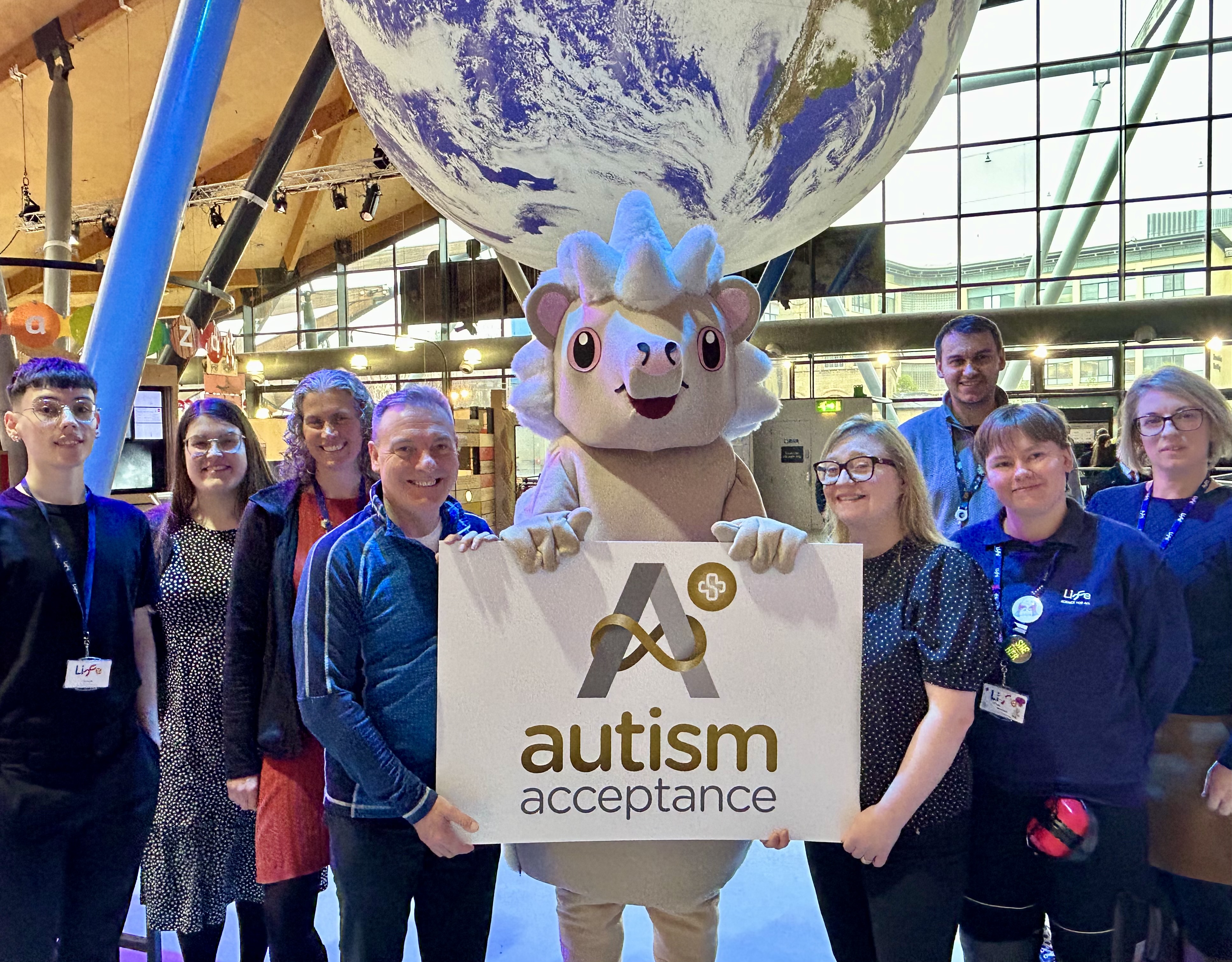 Group of people stood around a mascot holding the Autism Acceptance Plus Award inside the Life Science Centre