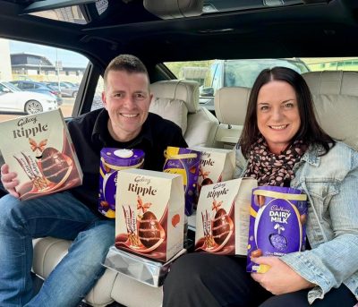 Two people in chauffeur vehicle holding lots of boxes of Easter Eggs and smiling to the camera.