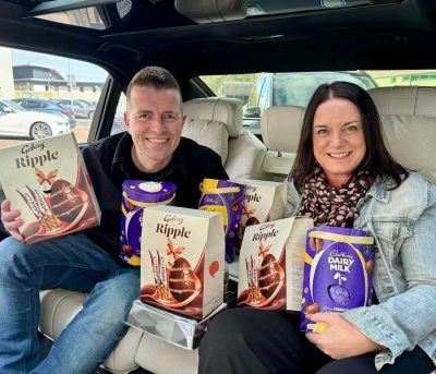 Two people in chauffeur vehicle holding lots of boxes of Easter Eggs and smiling to the camera.