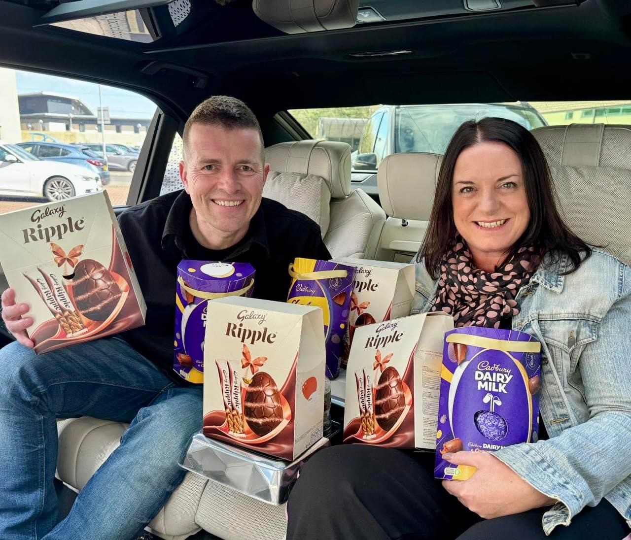 Two people in chauffeur vehicle holding lots of boxes of Easter Eggs and smiling to the camera.