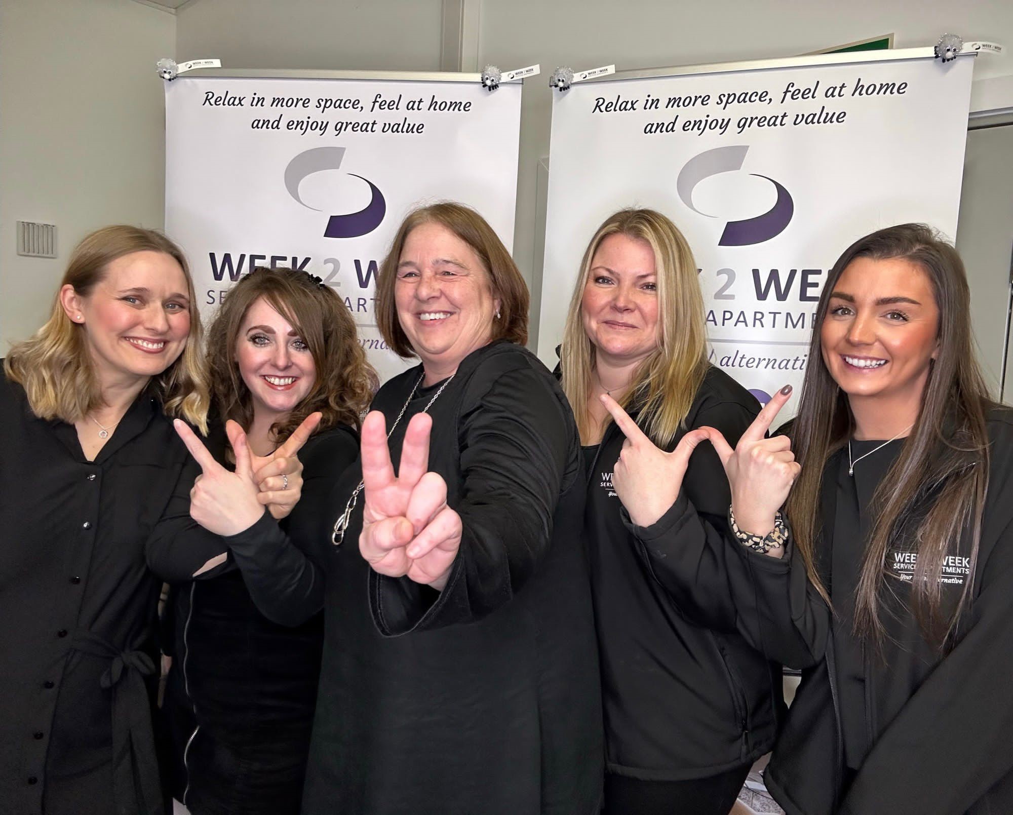 Week2Week team posing in front of branded banner stands and creating W2W symbols using their hands.