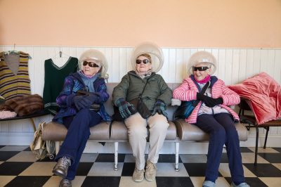 Three women sat under retro hair dryers with sunglasses on.