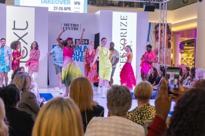 Models in brightly coloured clothing strutting down the runway with members of the audience looking on.