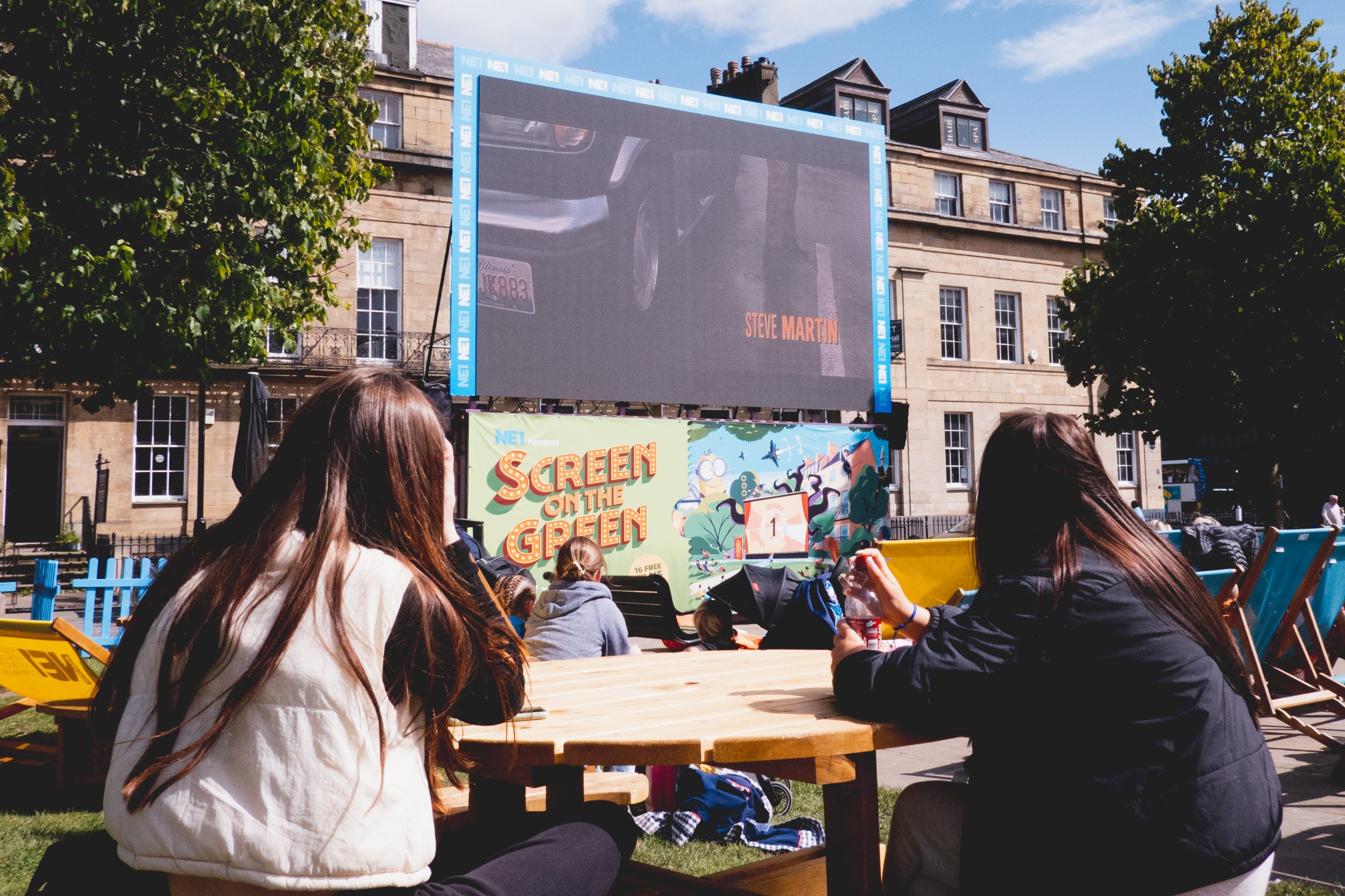 People sat on a picnic table looking up at the 'Screen on the Green' in the sunshine