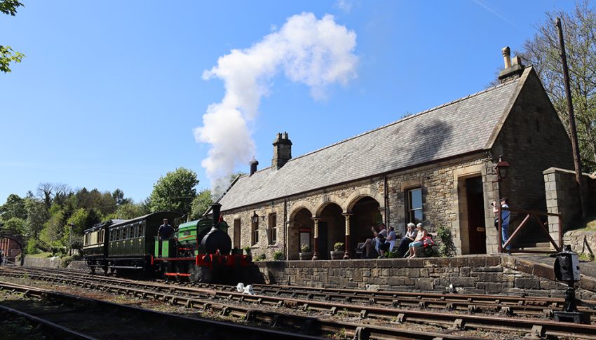 Green steam train on tracks outside of a stone built station.