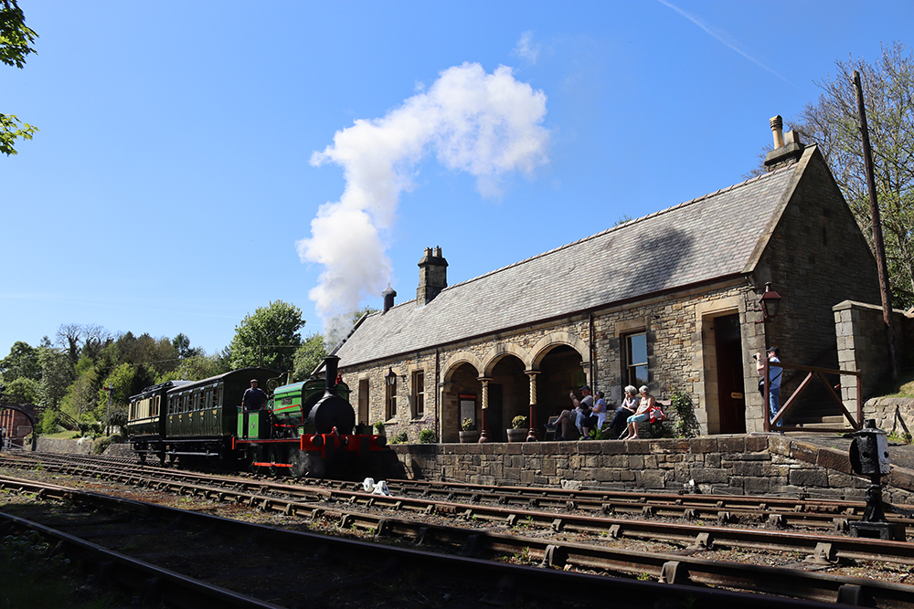 Green steam train on tracks outside of a stone built station.