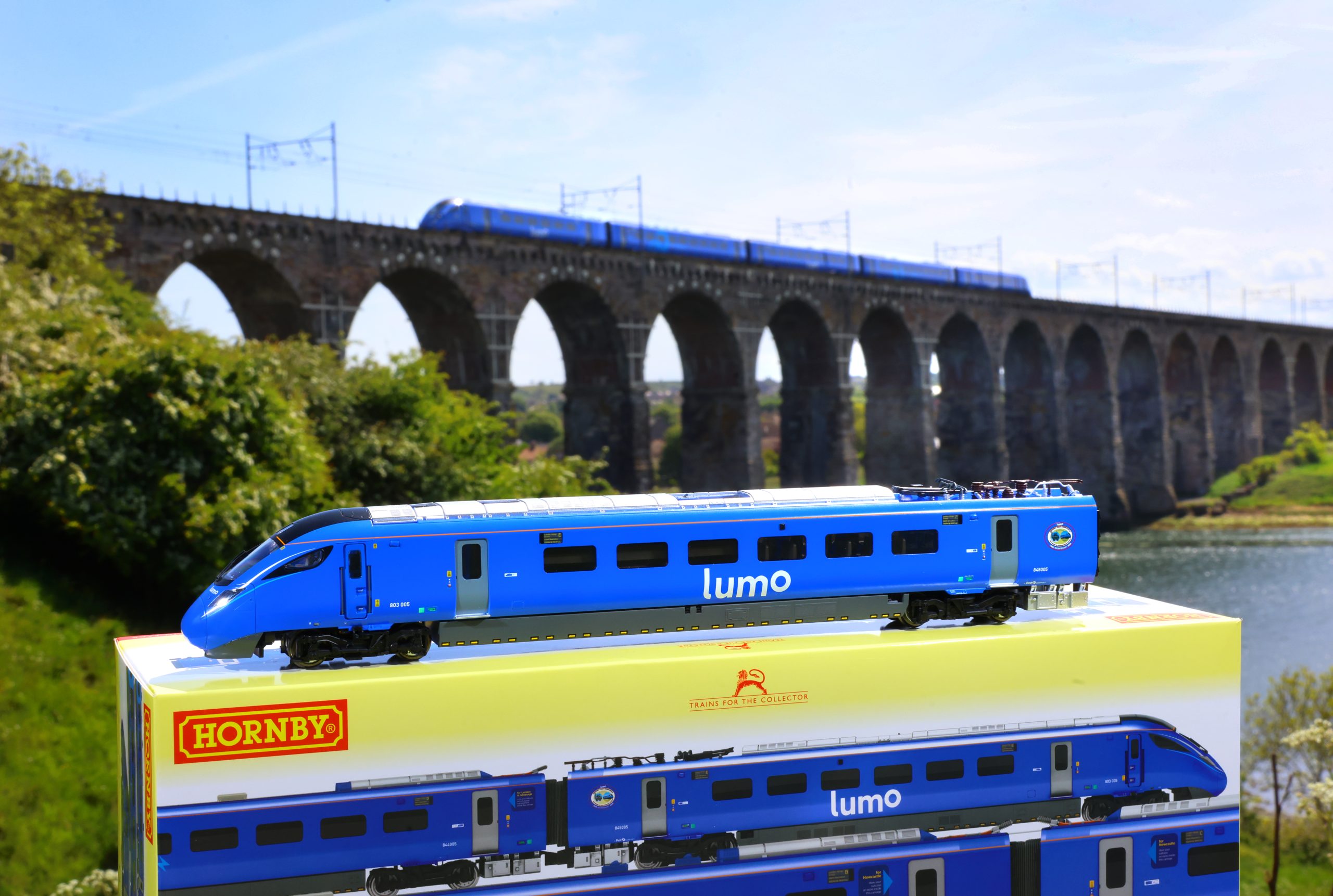 Lumo train model on top of its box underneath a rail bridge which has a real-life Lumo train crossing.
