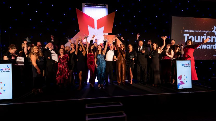 Large group of North East Tourism Awards winners stood on stage celebrating their awards in 'black tie' attire.