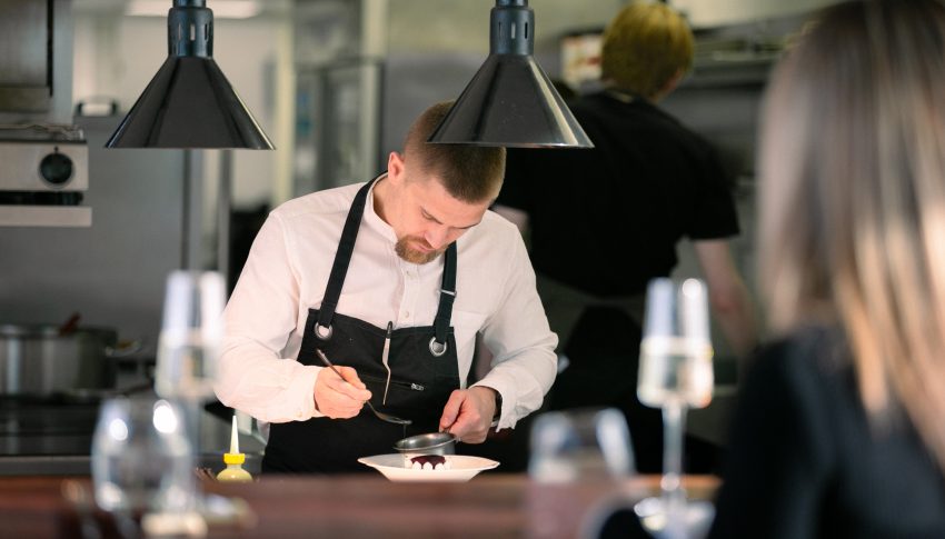 Chef in an open kitchen serving up a dessert in front of a guest.