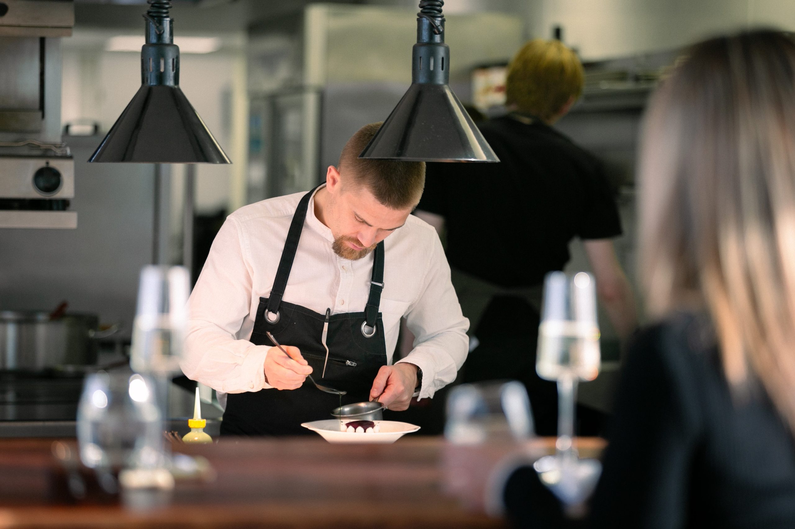 Chef in an open kitchen serving up a dessert in front of a guest.