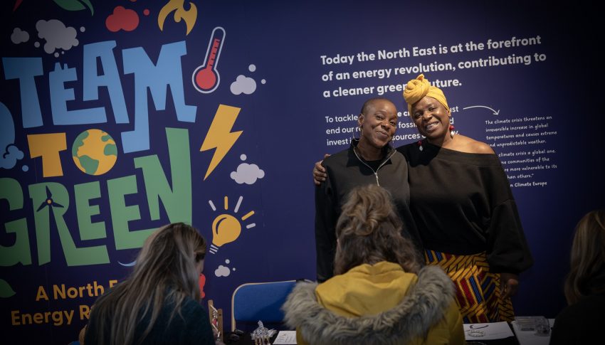 Two people stood together with arms around each other smiling to camera whilst others are sat in the foreground engaging in a creative activity.
