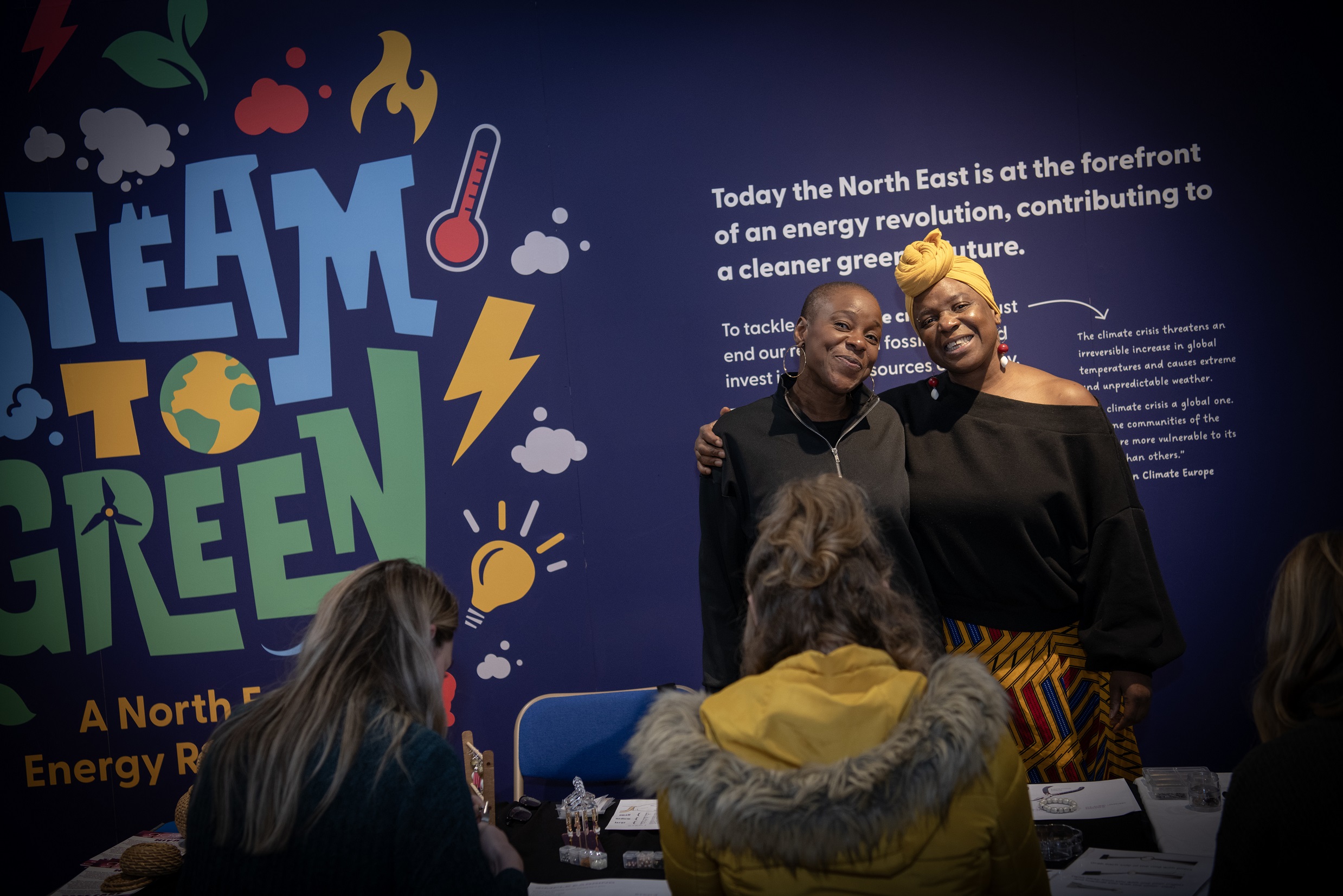 Two people stood together with arms around each other smiling to camera whilst others are sat in the foreground engaging in a creative activity.