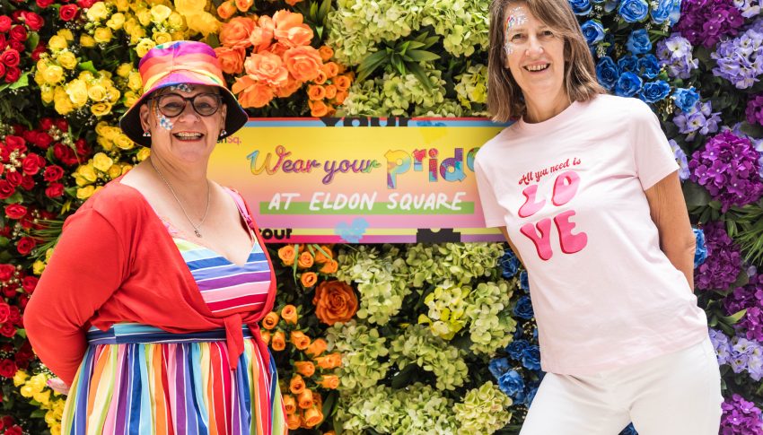 Two people wearing colourful Pride themed clothing stood in front of a rainbow floral backdrop.