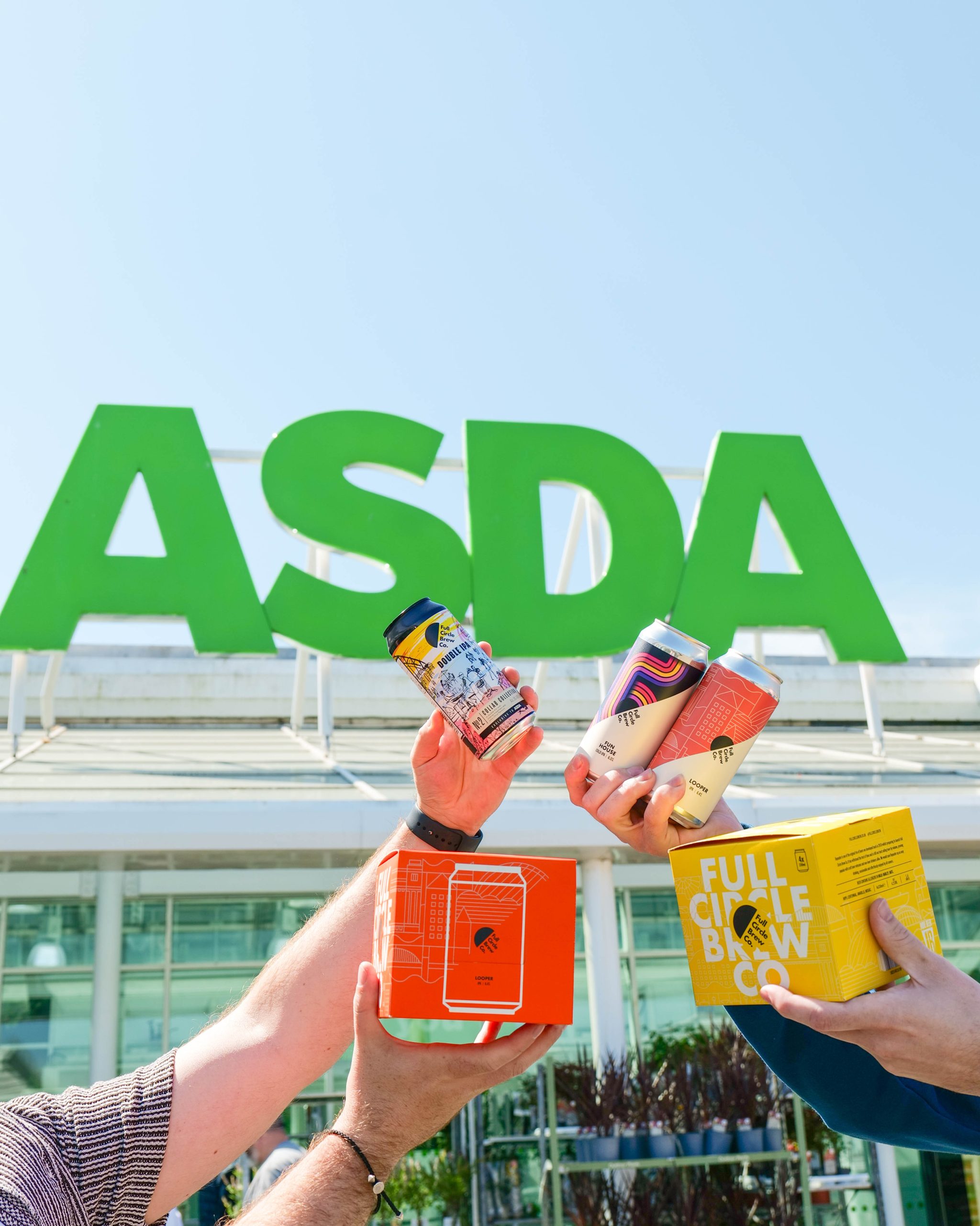 Colourful Full Circle Brew Co beer cans held up by hands in front of Asda sign