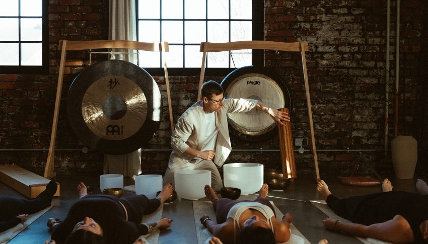 Leader of breathwork class sat at front of room surrounded by sound instruments and attendees lying on mats