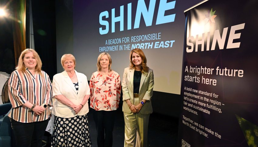 Group of women stood together on stage smiling to camera with the SHINE logo in the background on a large screen.