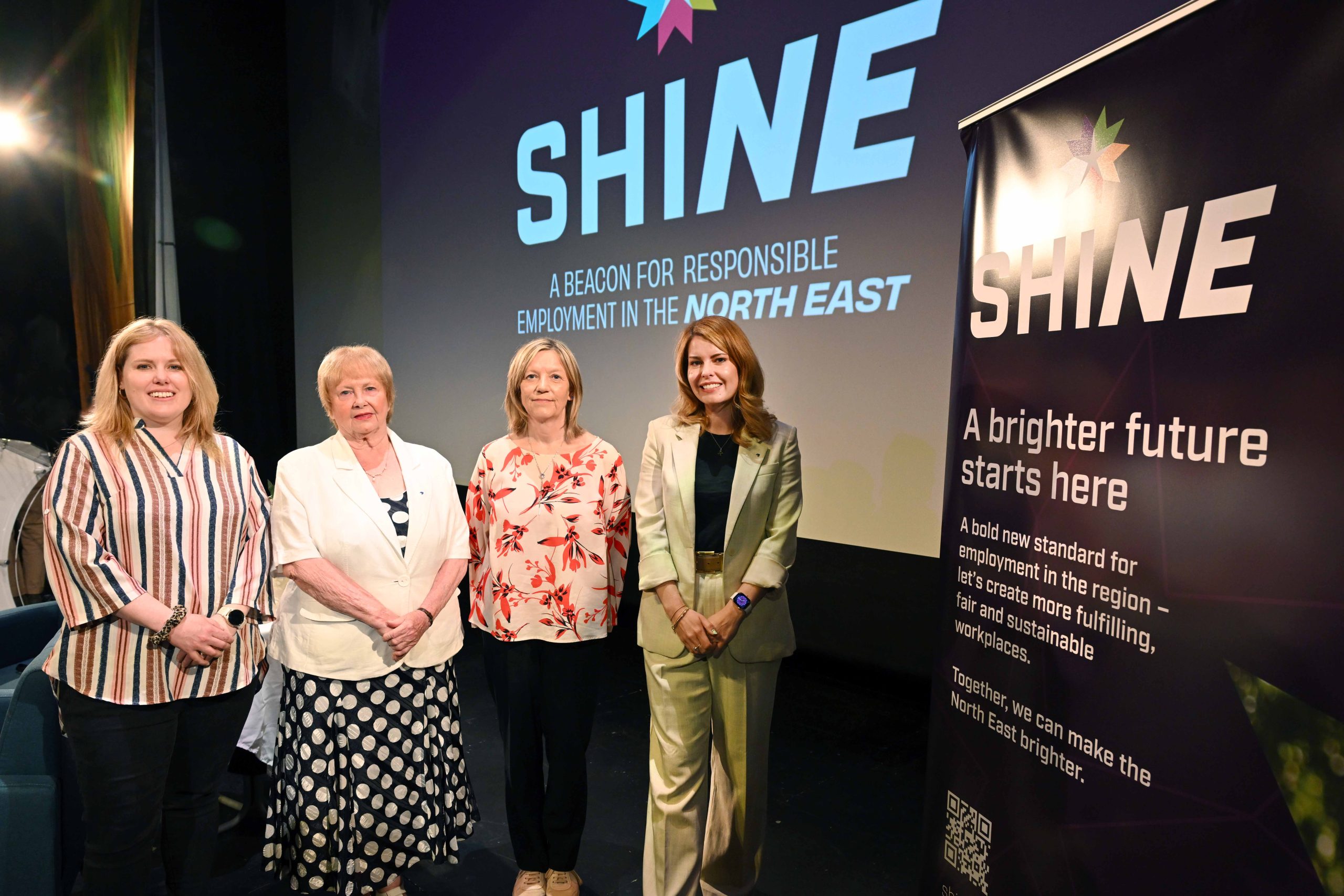 Group of women stood together on stage smiling to camera with the SHINE logo in the background on a large screen.