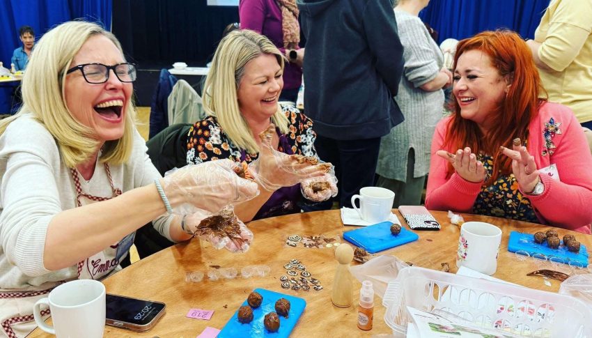 Three people sat around a table making chocolates and laughing hard.