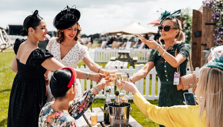 Women dressed in fancy dresses and fascinators cheers their drinks next to a racecourse.