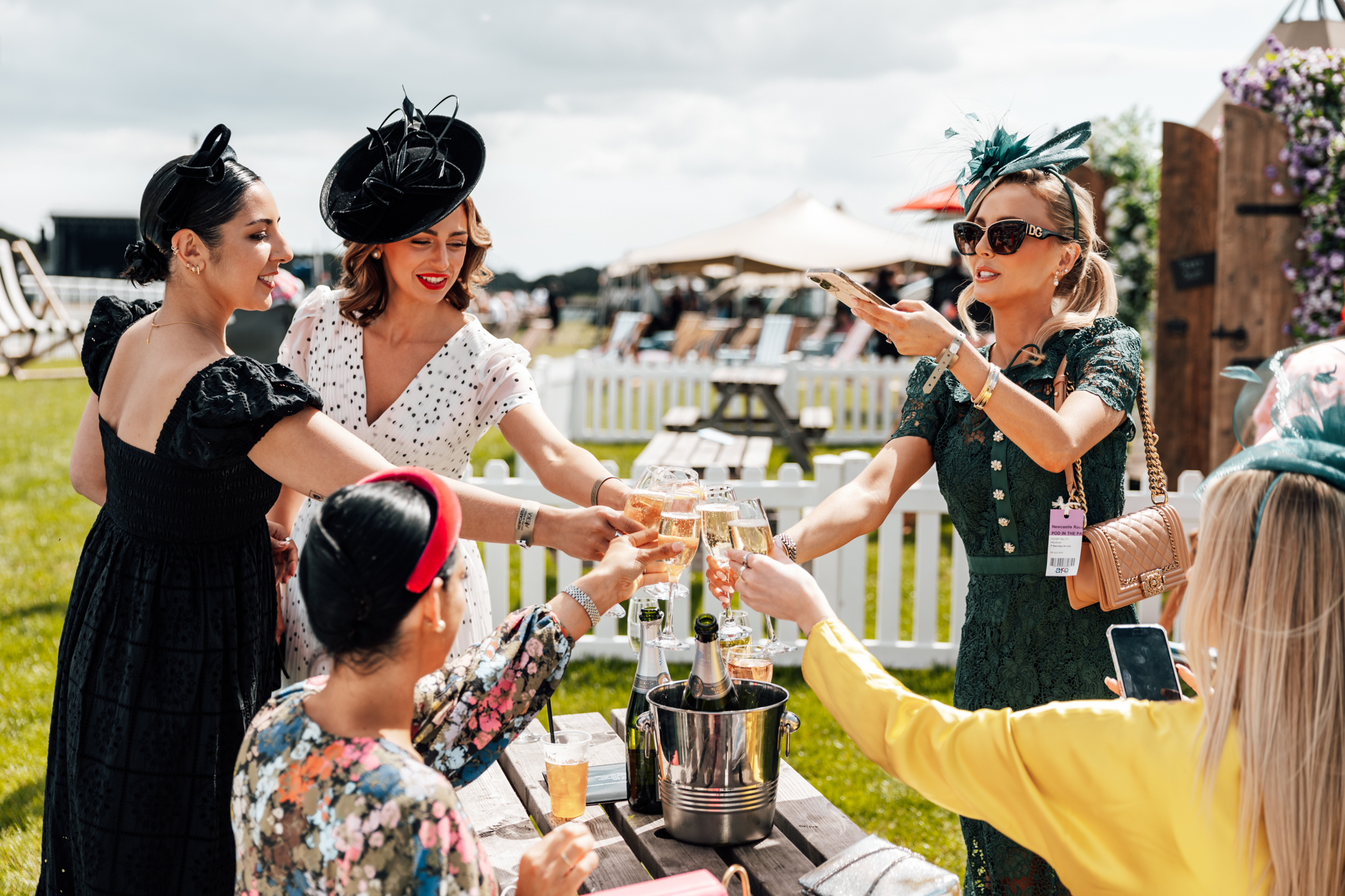Women dressed in fancy dresses and fascinators cheers their drinks next to a racecourse.