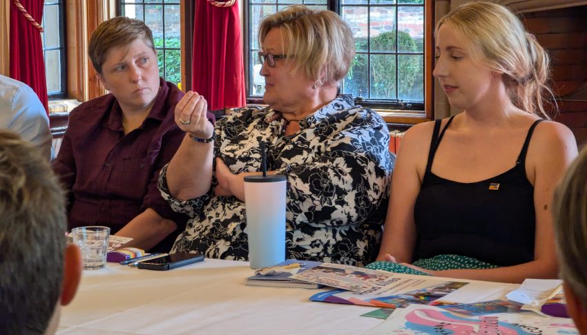 Person sat around table speaking as those sitting beside them listen intently.