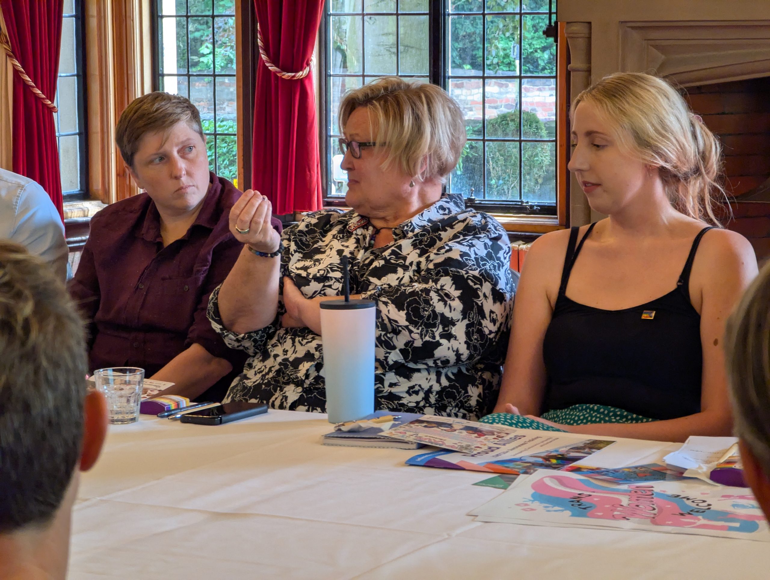 Person sat around table speaking as those sitting beside them listen intently.