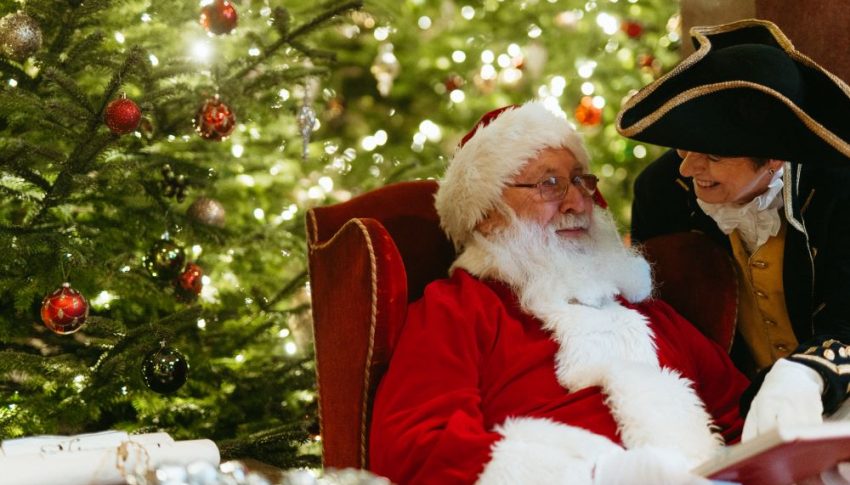 Older person dressed as Father Christmas sat in a red velvet armchair with Christmas tree in the background and person dressed in late 1700s costume bending down to talk to them.