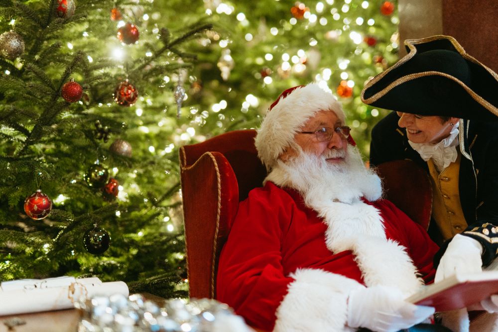 Older person dressed as Father Christmas sat in a red velvet armchair with Christmas tree in the background and person dressed in late 1700s costume bending down to talk to them.