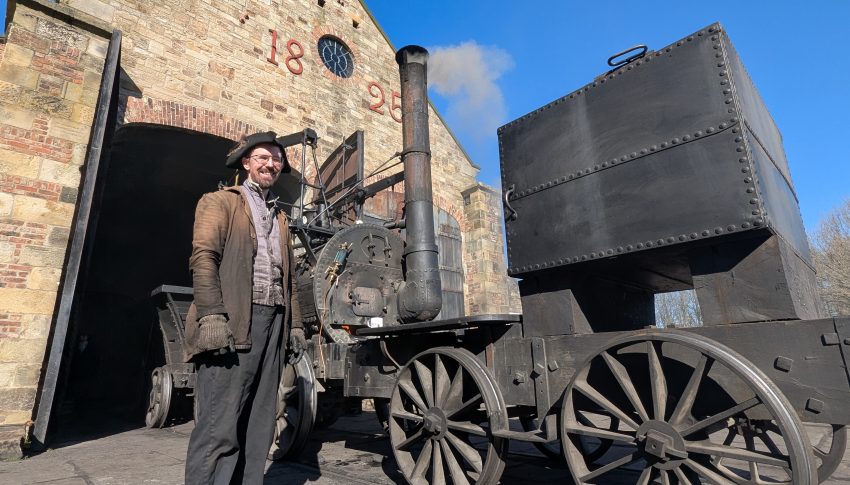 Beamish worker stood next to steam engine wearing heavy duty clothing which has soot on it.