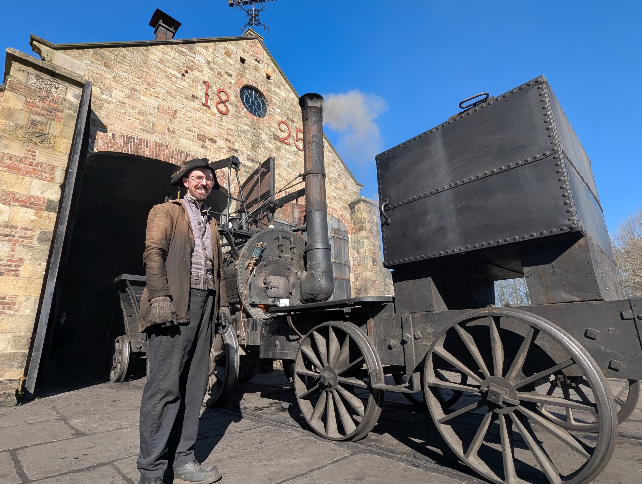 Beamish worker stood next to steam engine wearing heavy duty clothing which has soot on it.