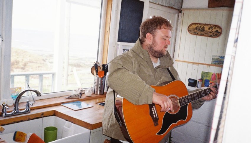Person stood in a kitchen strumming on an acoustic guitar