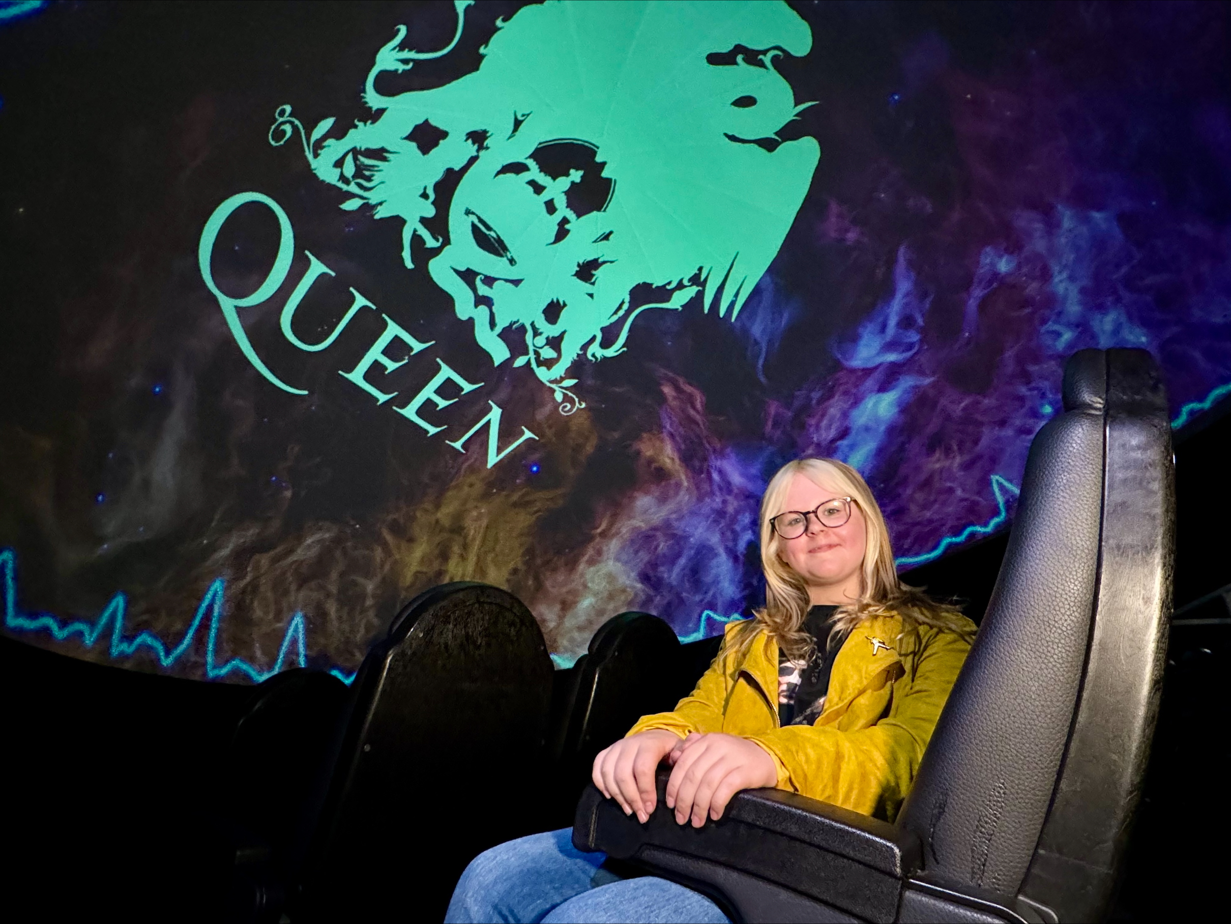 Ruby sat in cinema seat at Centre for Life with 'Queen' logo projected above her.