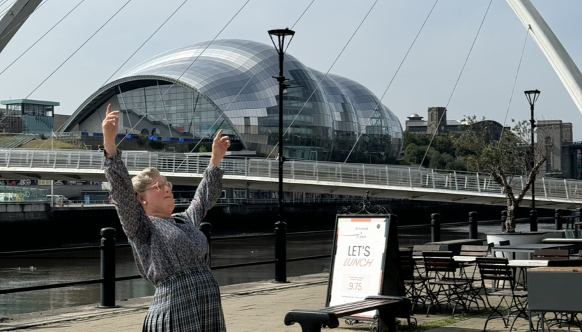 Person dressed as Mrs Doubtfire stood next to the Millennium Bridge reaching out their arms above their head.