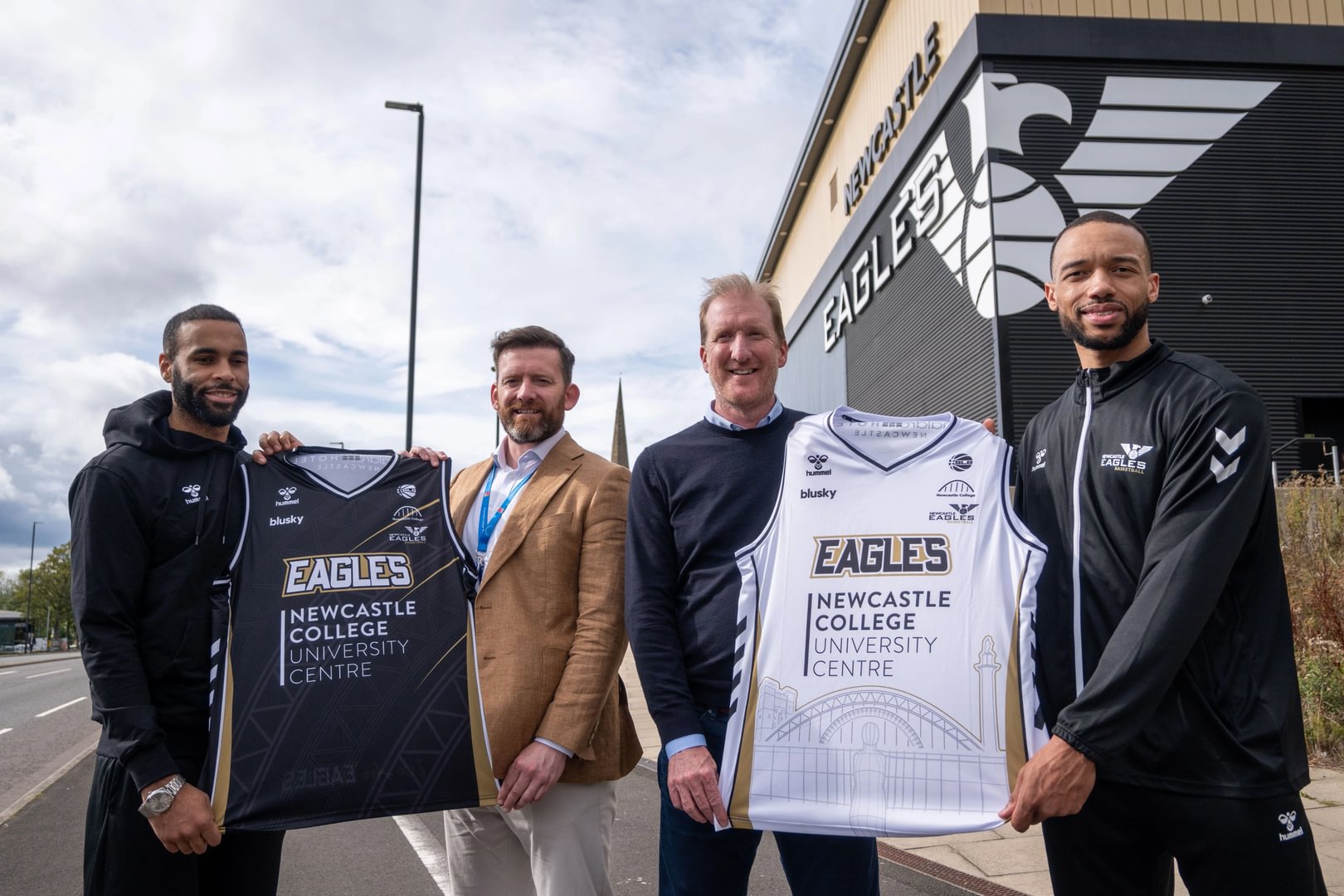 Newcastle Eagles Player Josh Ward Hibbert with Newcastle College Deputy Principal Alan Fenton, Eagles Managing Director Paul Blake and Player Marco Anthony holding the home and away basketball jerseys outside the Eagles stadium.
