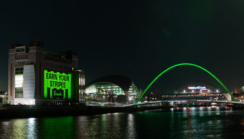 Quayside at nighttime with Millennium Bridge lit up in 'Fenwick green' and Baltic gallery wall facing the Tyne lit up and reading 'Earn your Stripes'