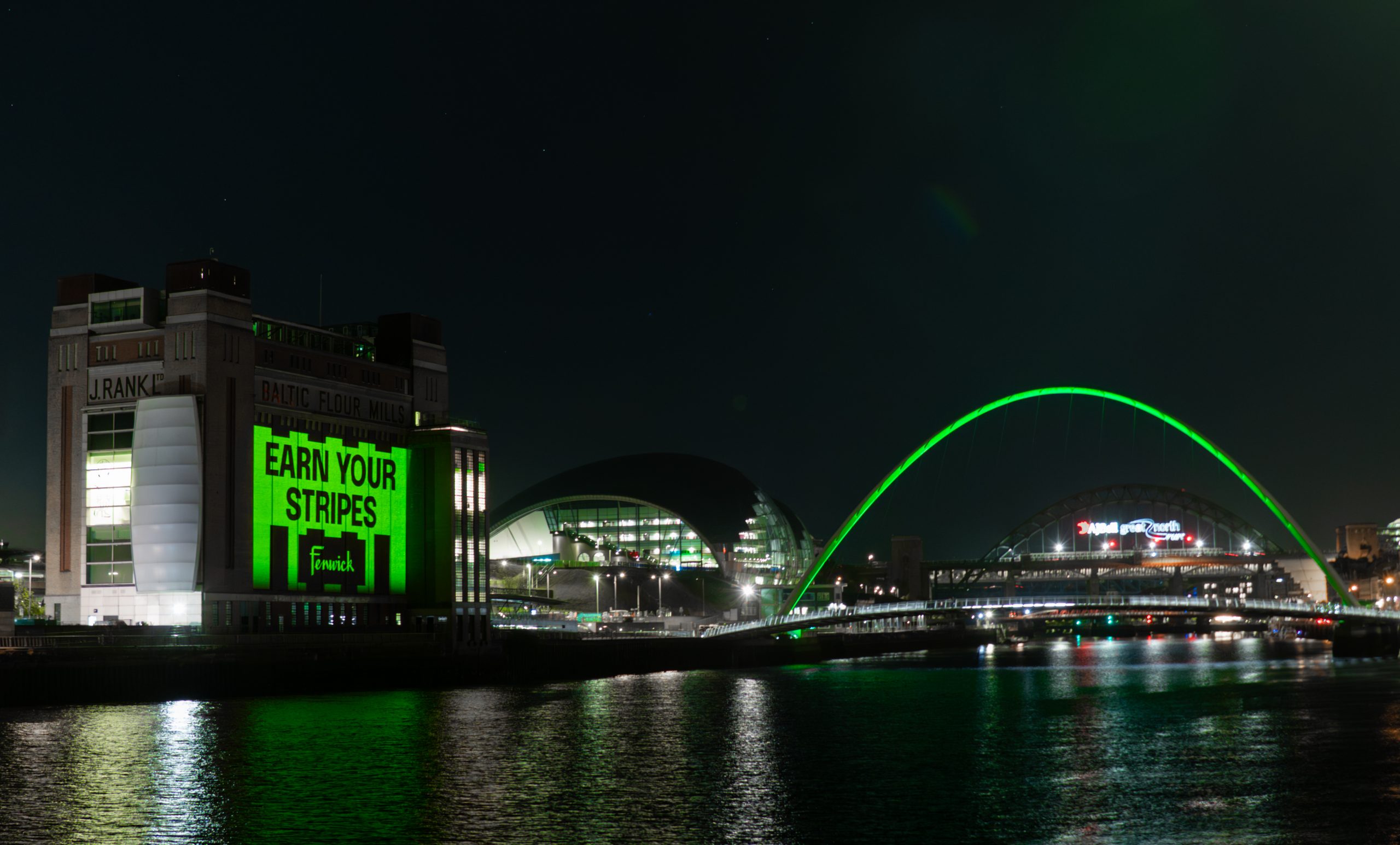 Quayside at nighttime with Millennium Bridge lit up in 'Fenwick green' and Baltic gallery wall facing the Tyne lit up and reading 'Earn your Stripes'