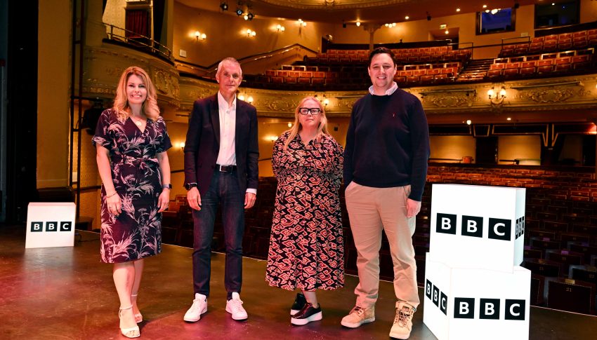 North East Screen Chief Executive, Alison Gwynn with North East Mayor, Kim McGuiness, Tees Valley Mayor, Ben Houchen, and BBC Director General, Tim Davie. They are all stood on stage in a theatre with two light-up BBC branded boxes to their right.
