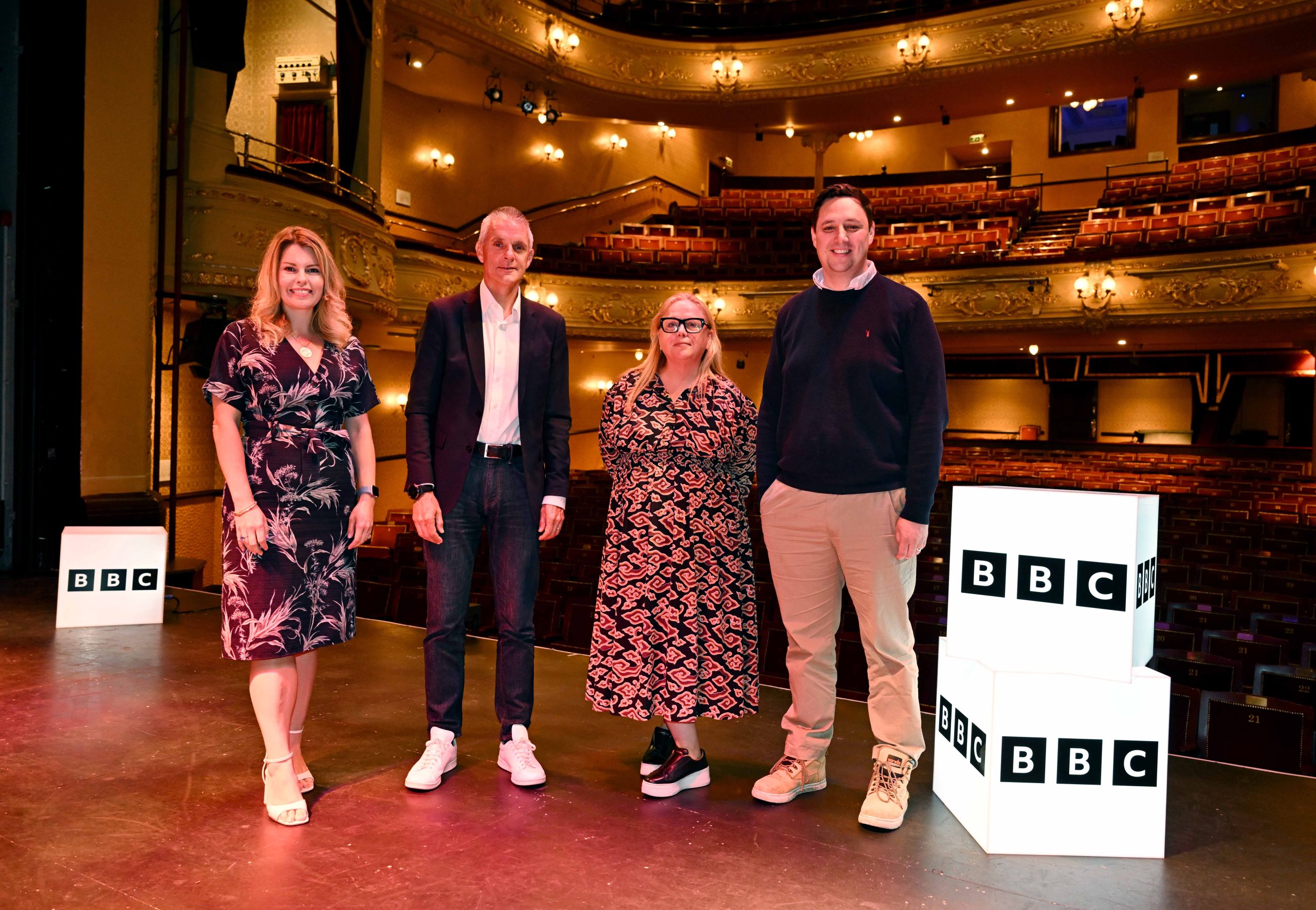 North East Screen Chief Executive, Alison Gwynn with North East Mayor, Kim McGuiness, Tees Valley Mayor, Ben Houchen, and BBC Director General, Tim Davie. They are all stood on stage in a theatre with two light-up BBC branded boxes to their right.