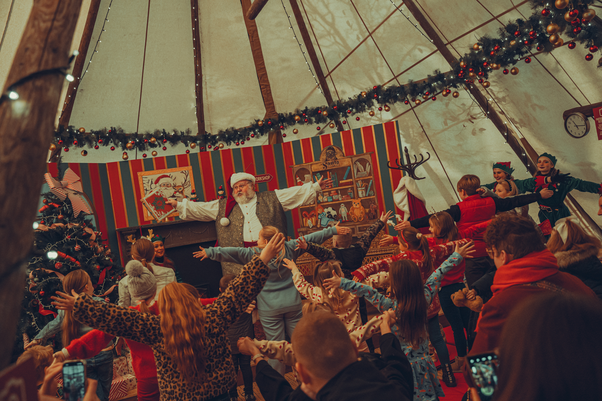 Person dressed as Santa Claus leading a dance inside a Christmas themed tipi with children following along in the foregound.
