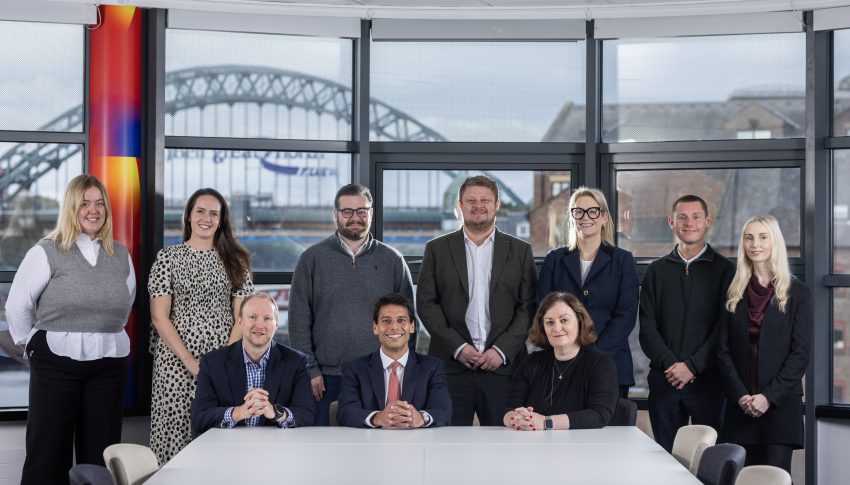 Group of new Ward Hadaway hires stood and sat in the boardroom which looks out to the River Tyne and the Tyne Bridge is in the background.