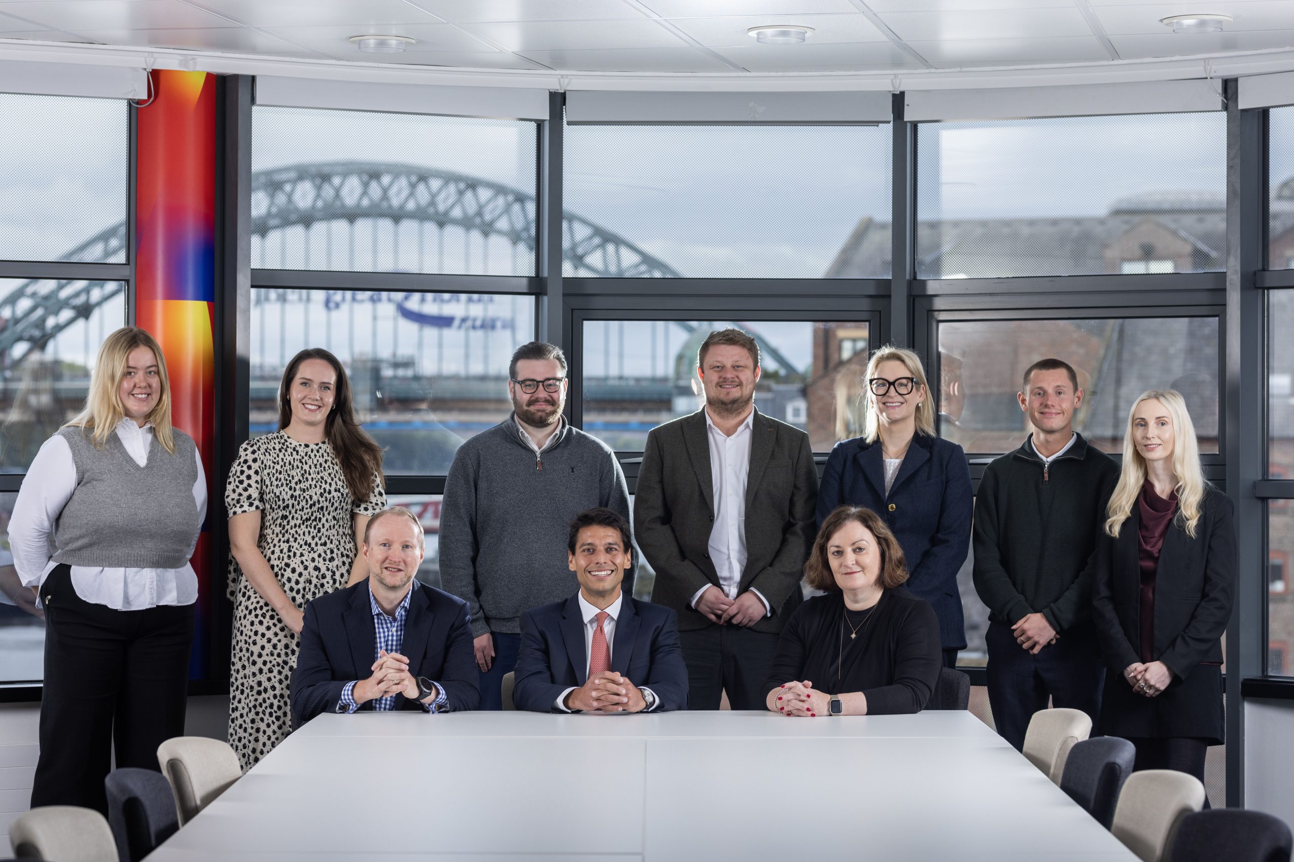 Group of new Ward Hadaway hires stood and sat in the boardroom which looks out to the River Tyne and the Tyne Bridge is in the background.