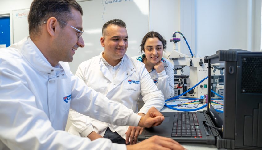 Three people in Newcastle University lab coats looking at a computer screen smiling, one is controlling the mouse.