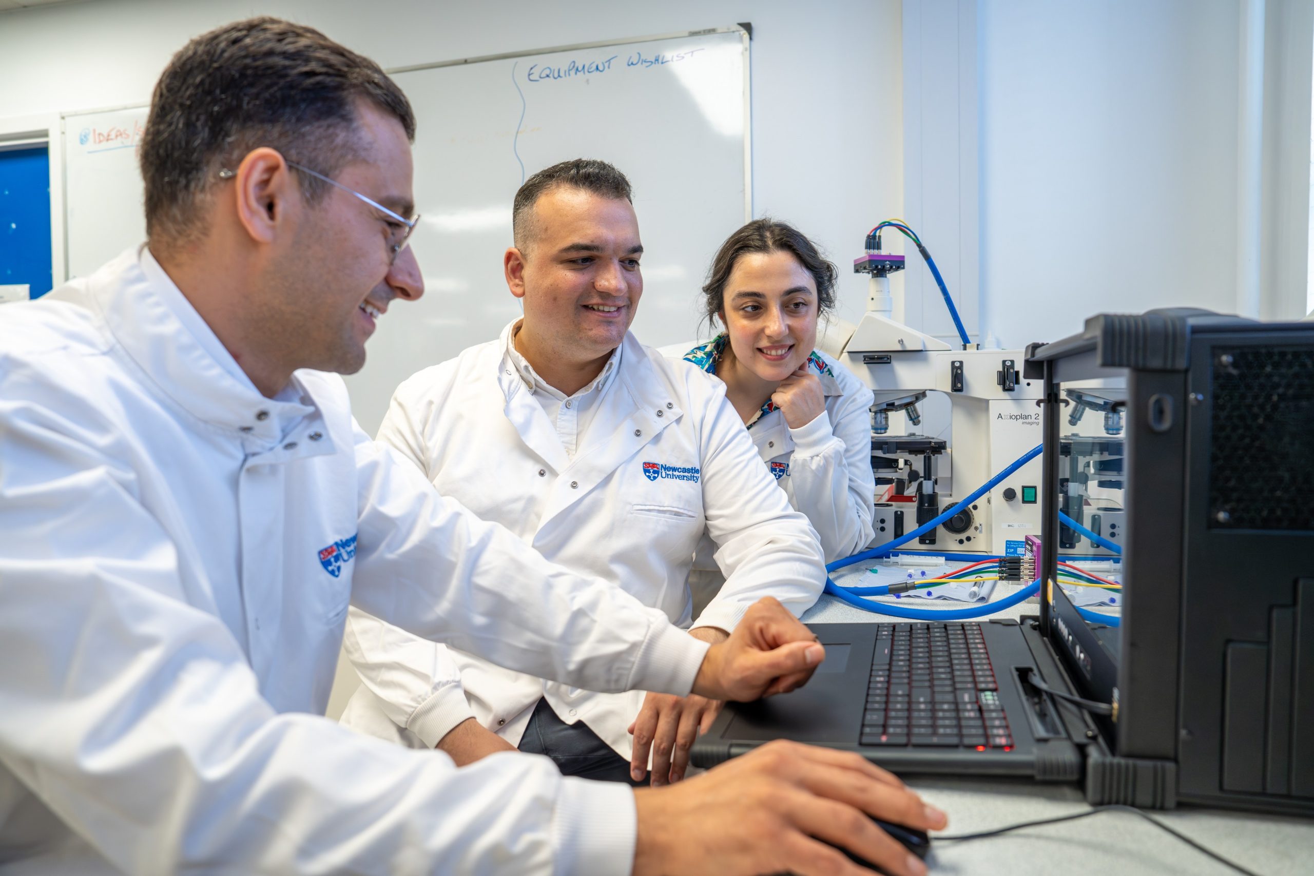 Three people in Newcastle University lab coats looking at a computer screen smiling, one is controlling the mouse.
