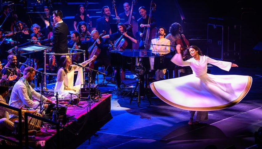 Orchestra and band performing on stage with person in a white dress dancing at the front, the skirt of the dress is flared out as they move in a circle.