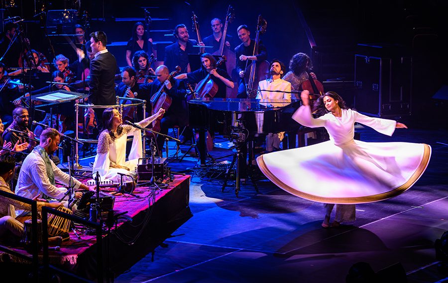 Orchestra and band performing on stage with person in a white dress dancing at the front, the skirt of the dress is flared out as they move in a circle.