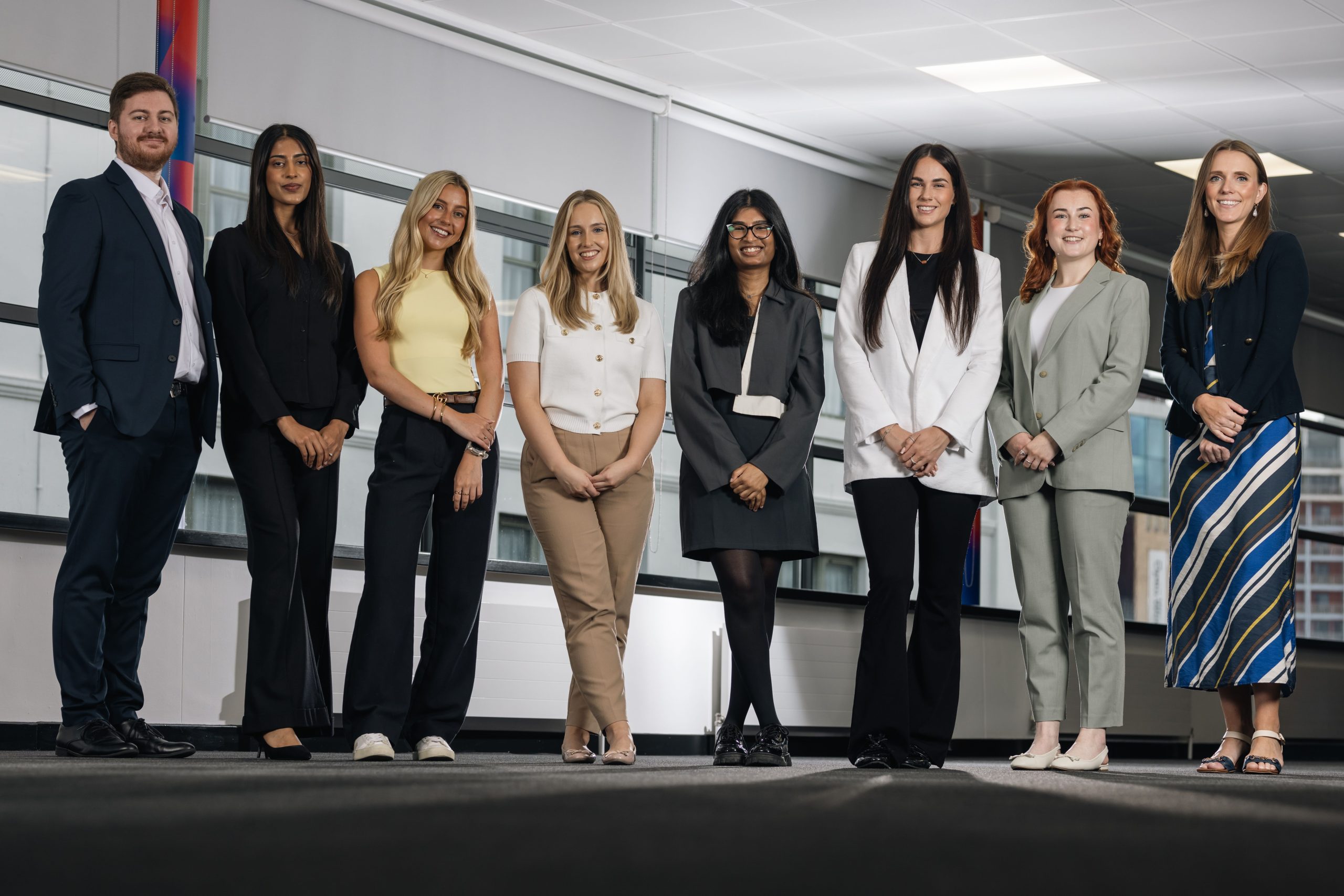 8 young people dressed smartly stood in a line smiling to camera in a plain looking room.