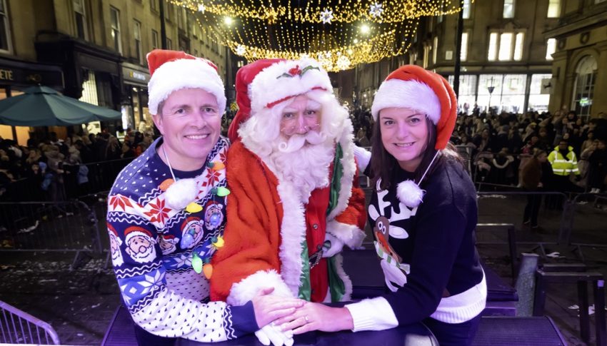 Steve, Karen and person dressed as Santa Claus pressing the button to turn on the Newcastle Christmas lights. Both Steve and Karen are wearing Christmas jumpers and Santa hats.