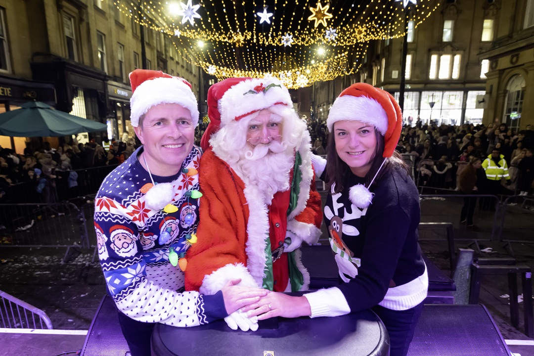 Steve, Karen and person dressed as Santa Claus pressing the button to turn on the Newcastle Christmas lights. Both Steve and Karen are wearing Christmas jumpers and Santa hats.